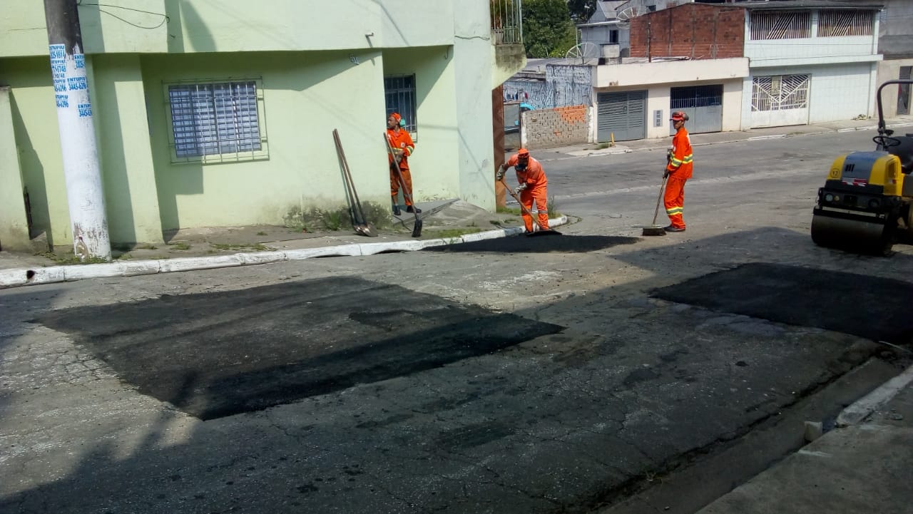 Três funcionários finalizam o asfaltamento de buraco em via.